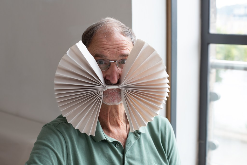 A white man with glasses holding a folded piece of paper in his mouth, which spans outwards like a giant bow-tie.
