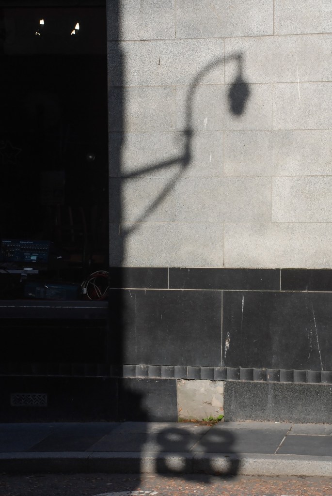 Shadows- of a lampost and a Co-op sign, fall on a building and pavement. 