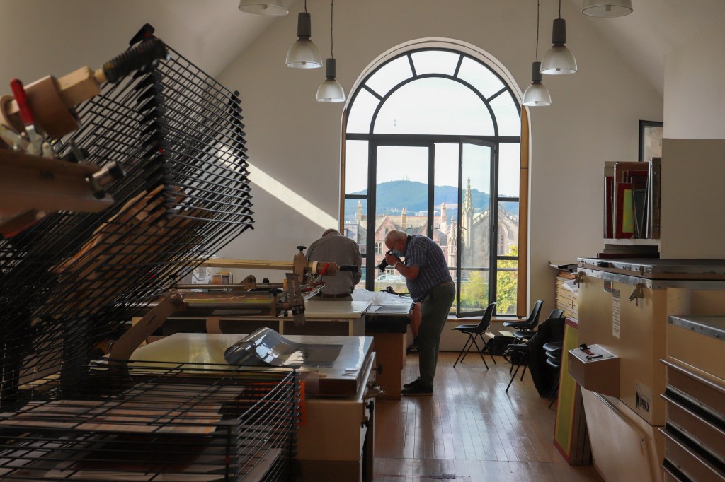 In the Highland Print Studio, amidst printing equipment. two men take photos of objects on a table. They are in front of a big window, which looks out onto old buildings across the river.