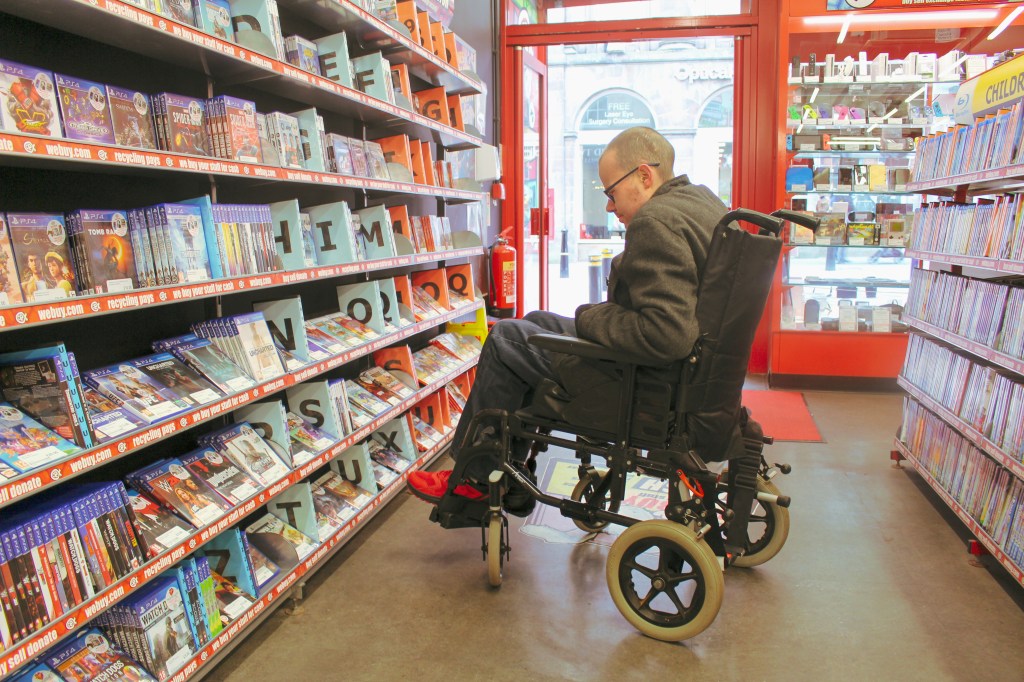 Brandon sits in his wheelchair, looks at shelves full of DVDs in a shop. Image by Evija Laivina. 